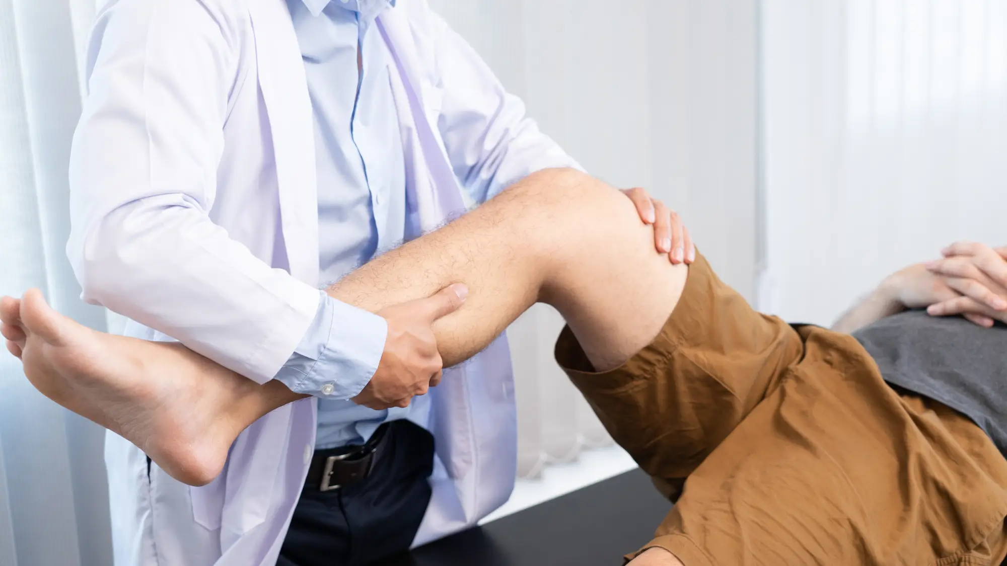 A healthcare professional in a white coat is assisting a patient lying on an examination table. The professional is holding and examining the patient's bent knee and calf, suggesting a physical evaluation or treatment session.