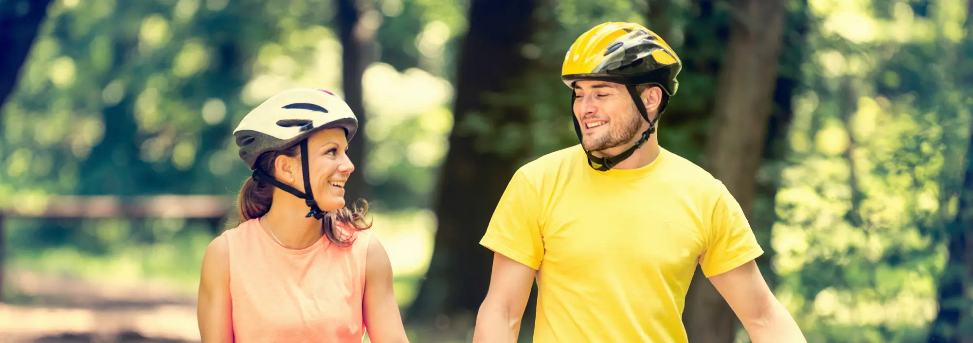 A woman and a man are smiling at each other as they walk with their bicycles. Both are wearing helmets; the woman is in a white helmet and pink top, and the man in a yellow helmet and matching yellow top. They are in a wooded park with green trees in the background.