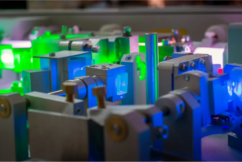 A close-up view of a laser laboratory setup with various high-tech equipment. The intricate arrangement of metallic components is illuminated by neon-green and blue lights, creating a futuristic atmosphere.