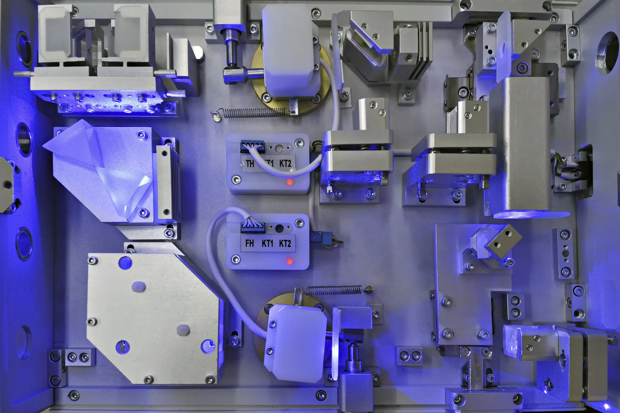Close-up of a sophisticated, modular machine component setup with metal parts, connectors, and tubing arranged on a mounting plate. The machine is illuminated by blue lighting, highlighting its intricate mechanical elements.