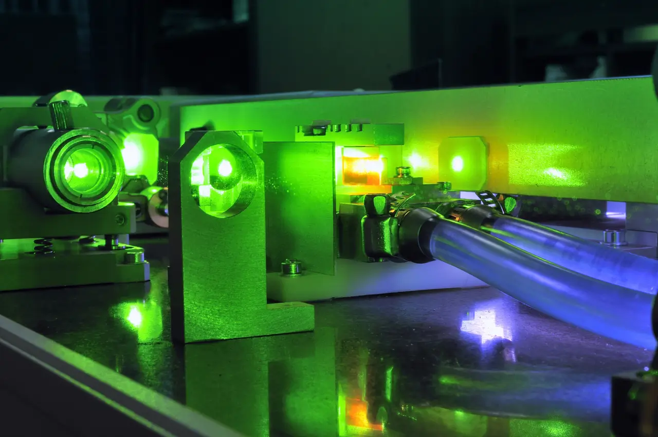 A close-up of a laser setup with green lights illuminating various components. Metallic tubes and reflective surfaces are visible, with a vibrant green glow highlighting the intricate details of the apparatus.
