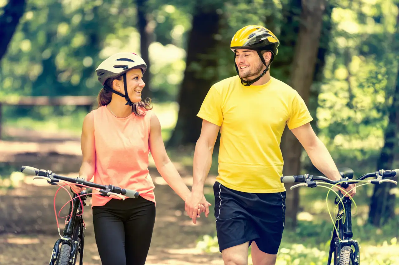 A woman and a man are smiling at each other as they walk with their bicycles. Both are wearing helmets; the woman is in a white helmet and pink top, and the man in a yellow helmet and matching yellow top. They are in a wooded park with green trees in the background.